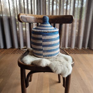 May include: A blue and white striped woven basket with a lid, sitting on a chair with a faux fur throw. The basket is made of natural materials and has a rustic, handmade look.