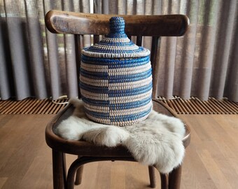 May include: A blue and white striped woven basket with a lid, sitting on a chair with a faux fur throw. The basket is made of natural materials and has a rustic, handmade look.