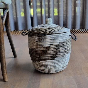 Puede incluir: Una cesta de almacenamiento redonda y tejida que cuenta con una tapa con un pequeño pomo y dos asas laterales. La cesta está fabricada con fibras naturales en un patrón de bandas horizontales alternas en colores blanco, beige y marrón oscuro.