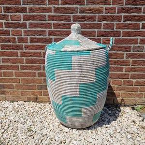 Puede incluir: Una cesta grande, tejida, con tapa. La cesta presenta un patrón geométrico en color turquesa y blanco roto. Tiene forma redondeada y dos asas pequeñas. El fondo es una pared de ladrillo y pequeñas piedras blancas.