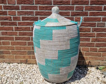May include: A large, woven basket with a lid. The basket features a geometric pattern in teal and off-white. It has a rounded shape and two small handles. The background is a brick wall and small white stones.