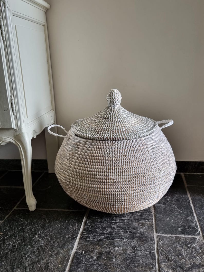 May include: A round, white woven basket with a matching conical lid and two side handles. The basket features a textured, coiled construction and a pointed finial on top of the lid, providing a structured storage solution with a neutral aesthetic.