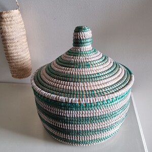 May include: A round woven basket with a lid, featuring a spiral design in green, white, and natural tones. The basket is on a white surface, with a smaller woven item hanging in the background. The basket is likely used for storage or decoration.
