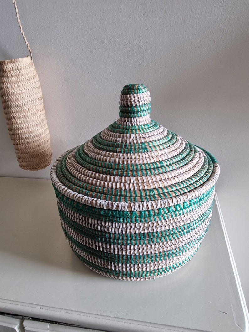 May include: A round woven basket with a lid, featuring a spiral design in green, white, and natural tones. The basket is on a white surface, with a smaller woven item hanging in the background. The basket is likely used for storage or decoration.