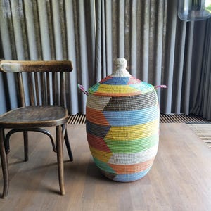 Op de afbeelding: Een hoge, cilindrische gevlochten mand met een conisch deksel en twee handvatten aan de zijkant. De mand heeft een geometrisch patroon in levendige kleuren zoals geel, oranje, groen, blauw en wit, gemaakt van strak geweven, getextureerd materiaal.