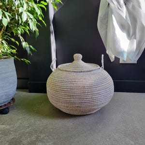 May include: A round, woven storage basket with a matching lid and two white rope handles. The basket features a textured, light-colored weave pattern and a rounded, bulbous shape, providing a functional container for organizing household items.