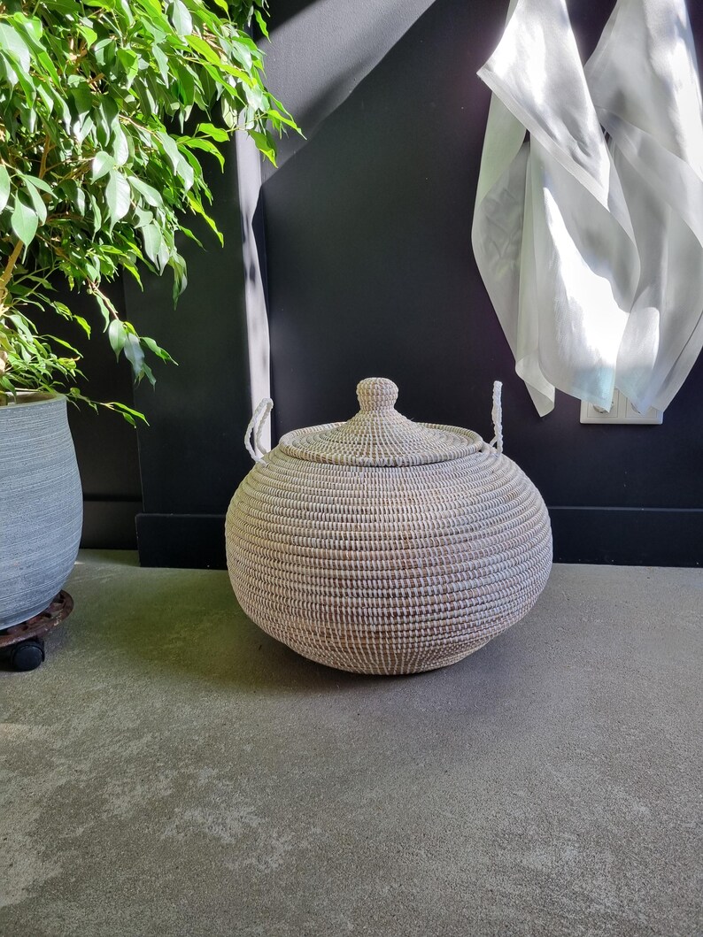 May include: A round, woven storage basket with a matching lid and two white rope handles. The basket features a textured, light-colored weave pattern and a rounded, bulbous shape, providing a functional container for organizing household items.