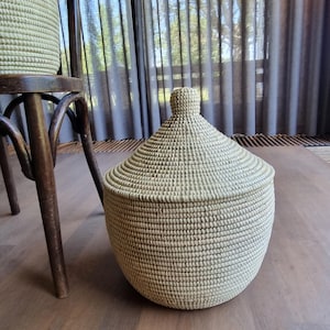 May include: A round, light-colored woven basket with a matching conical lid featuring a small knob handle at the top. The basket has a textured, coiled construction and a wide, bulbous body, providing a sturdy container for storage and organization.