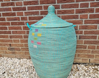 May include: A large, turquoise woven basket with a lid. The basket has a rounded shape and is made of tightly woven strands. The lid has a knob on top and small handles on the sides. The basket is decorated with colorful accents and sits in front of a brick wall.