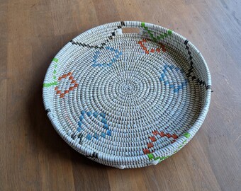 May include: Round woven basket with a handle, featuring a cream-colored base and colorful geometric patterns in blue, orange, and green. The basket is made of natural materials and has a circular design.
