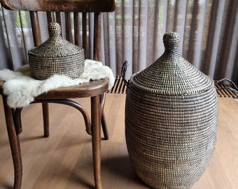 May include: Two black and white woven storage baskets with lids. The smaller basket is on a wooden chair, and the larger basket is on the floor.