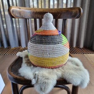 May include: A white woven basket with a colorful striped pattern sits on a furry white rug on a wooden chair. The basket has a round shape with a pointed top.