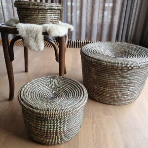 May include: Three woven baskets with lids in varying sizes. The baskets are black and beige, with a spiral design on the lids. One basket sits on a wooden chair with a fur throw, while the other two are on a wooden floor.
