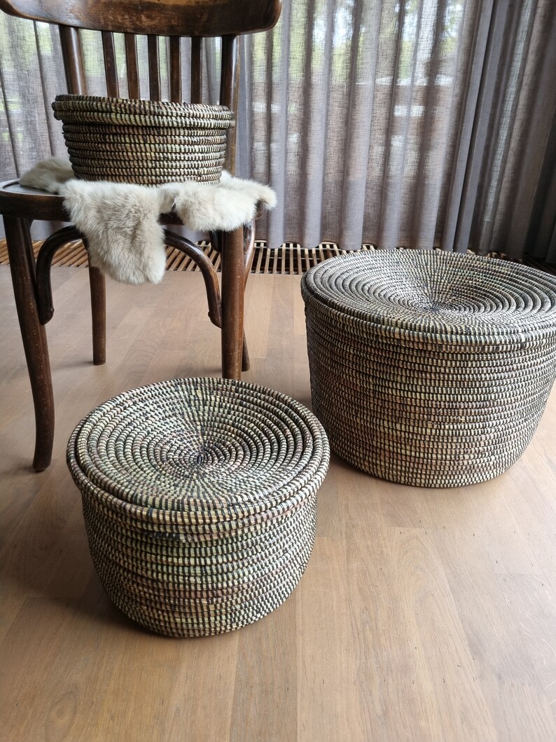 May include: Three woven baskets with lids in varying sizes. The baskets are black and beige, with a spiral design on the lids. One basket sits on a wooden chair with a fur throw, while the other two are on a wooden floor.