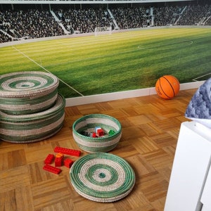 May include: A collection of woven baskets in varying sizes, featuring a green and natural color scheme. The baskets are arranged on a wooden floor, with an orange basketball and red toy blocks nearby. A large stadium mural is in the background.