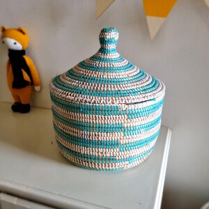 May include: A round, lidded basket with a turquoise and white striped design. The basket is made of woven material and sits on a white surface. A stuffed fox toy with a black scarf is in the background.