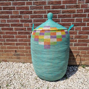 Puede incluir: Una gran cesta de almacenamiento tejida con tapa. La cesta es turquesa con un colorido patrón geométrico en la tapa. Tiene dos asas de cuerda azul y está colocada contra una pared de ladrillo.