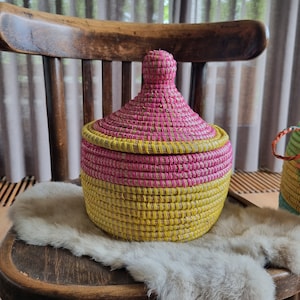 May include: A woven basket with a lid, featuring a pink and yellow colour scheme. The basket is sitting on a wooden chair with a white fur throw.