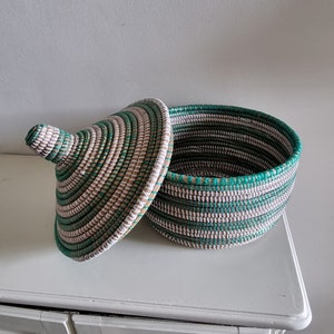 May include: A round, lidded basket with a woven design in shades of green, white, and tan. The lid is slightly ajar, revealing the interior. The basket is sitting on a white surface.