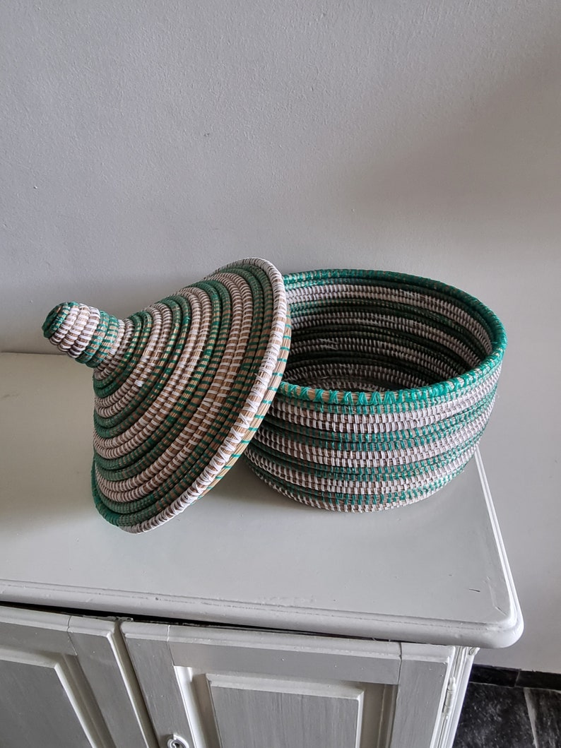 May include: A round, lidded basket with a woven design in shades of green, white, and tan. The lid is slightly ajar, revealing the interior. The basket is sitting on a white surface.
