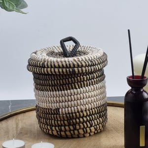May include: A woven basket with a lid, featuring alternating bands of black, white, and natural fibers. The basket sits on a wooden tray, with a dark red diffuser bottle and two small white candles nearby. The lid has a black handle.