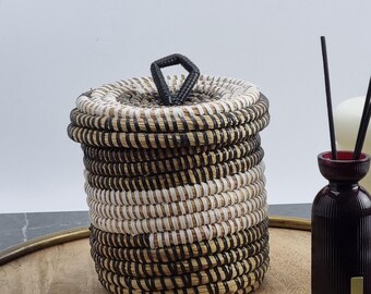 May include: A woven basket with a lid, featuring alternating bands of black, white, and natural fibers. The basket sits on a wooden tray, with a dark red diffuser bottle and two small white candles nearby. The lid has a black handle.