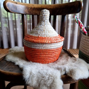 Op de afbeelding: Een geweven mand met een wit en oranje ontwerp staat op een donzige ondergrond op een houten stoel. De mand heeft een ronde, bolle vorm met een smalle hals.