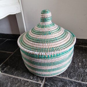 May include: A round, lidded basket with a woven design. The basket features alternating bands of natural, white, and green fibers. The lid has a conical shape with a small handle on top. The basket is sitting on a dark tiled surface.