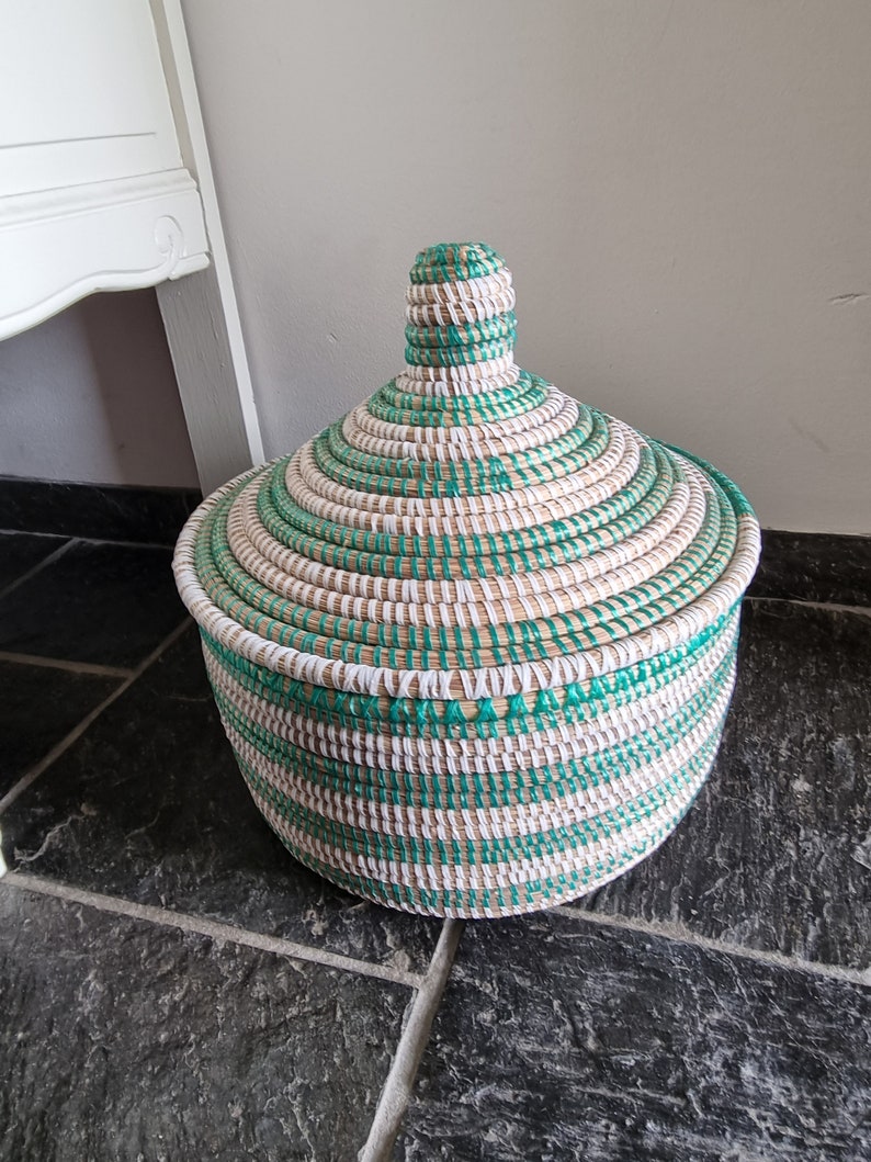May include: A round, lidded basket with a woven design. The basket features alternating bands of natural, white, and green fibers. The lid has a conical shape with a small handle on top. The basket is sitting on a dark tiled surface.