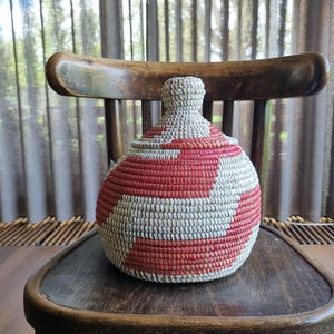 May include: A round, lidded basket featuring a woven pattern of natural beige and vibrant red fibers. The container has a bulbous body and a tapered, dome-shaped lid with a small, rounded knob at the top, constructed from coiled plant material.