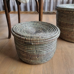 Puede incluir: Una cesta redonda tejida con tapa, con un diseño en espiral en tonos naturales y negros. La cesta está hecha de fibras tejidas apretadamente, creando una superficie texturizada. La tapa encaja perfectamente.