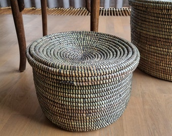 May include: A round, woven basket with a lid, featuring a spiral design in natural and black tones. The basket is made of tightly woven fibers, creating a textured surface. The lid fits snugly on top, and another similar basket is visible in the background.