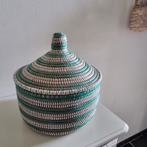 May include: A round, lidded basket with a woven design. The basket features alternating bands of teal, white, and natural-colored fibers. The lid has a conical shape, and the basket sits on a white surface.