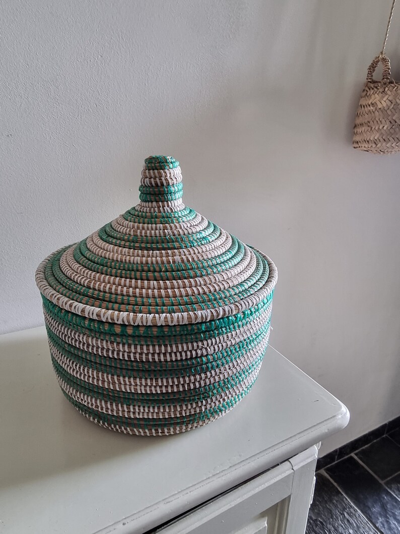 May include: A round, lidded basket with a woven design. The basket features alternating bands of teal, white, and natural-colored fibers. The lid has a conical shape, and the basket sits on a white surface.
