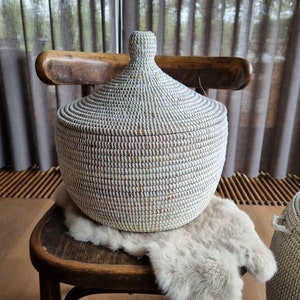 May include: White woven storage basket with a lid, sitting on a wooden chair with a faux fur throw. The basket has a round shape and a slightly domed lid.