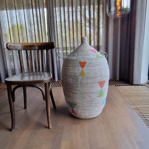 Op de afbeelding: Een grote, geweven mand met een conisch deksel, versierd met kleurrijke geometrische vormen. De mand is lichtgrijs en staat op een houten oppervlak. Links van de mand staat een houten stoel.