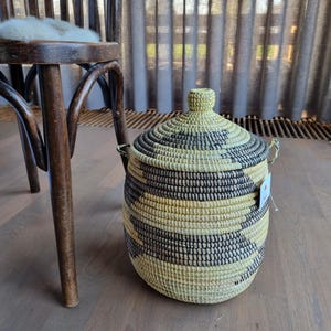May include: A large, round, woven storage basket with a lid. The basket is made of natural materials and has a black and white striped pattern. The basket is sitting on a wooden floor.