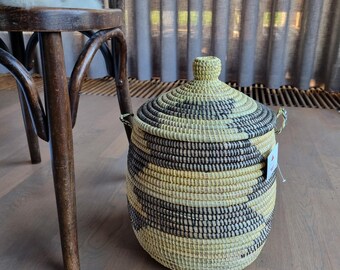 May include: A large, round, woven storage basket with a lid. The basket is made of natural materials and has a black and white striped pattern. The basket is sitting on a wooden floor.