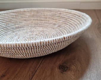May include: A woven, oval-shaped basket with a natural, light brown base and a white, textured rim. The basket is set on a wooden floor, showcasing its handcrafted design and neutral color palette.