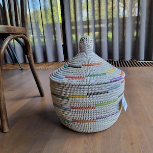 May include: A round, woven basket with a matching conical lid. The basket is primarily light beige with horizontal, intermittent dashes of colour including orange, yellow, green, blue, and purple. It features a small, rounded knob on the lid.