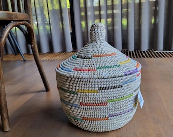 May include: A round, woven basket with a matching conical lid. The basket is primarily light beige with horizontal, intermittent dashes of color including orange, yellow, green, blue, and purple. It features a small, rounded knob on the lid.