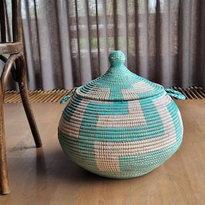 May include: A round, lidded basket with a turquoise and beige geometric pattern. The basket has a bulbous shape and two small handles. The lid has a rounded top. The basket is made of woven material and sits on a wooden floor.