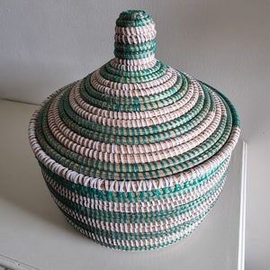 May include: A round, lidded basket with a woven design. The basket features alternating bands of natural, white, and teal-green fibers. The lid has a conical knob. The basket is sitting on a white surface.