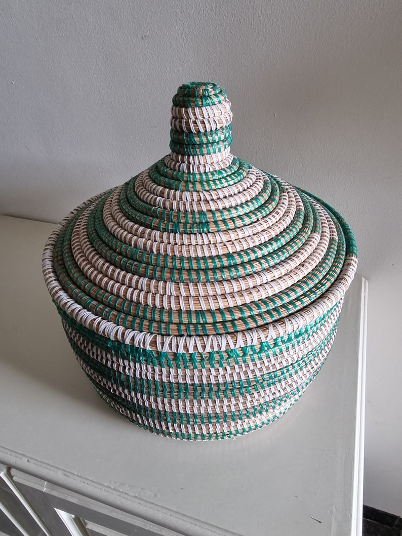 May include: A round, lidded basket with a woven design. The basket features alternating bands of natural, white, and teal-green fibers. The lid has a conical knob. The basket is sitting on a white surface.