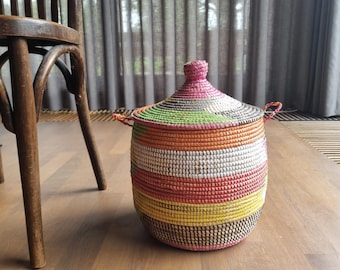 May include: A cylindrical woven basket with a conical lid and two small side handles. The basket features horizontal bands of vibrant colors including pink, orange, white, yellow, and dark brown, constructed from coiled material with a textured finish.