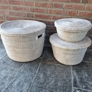May include: Three white wicker baskets with lids. The largest basket is on the left and the two smaller baskets are stacked on the right. The baskets are on a grey tiled floor.