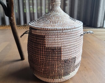 May include: A cylindrical woven basket with a matching lid and two side handles. The basket features a pattern of white, tan, and dark brown fibers coiled together. The lid has a small, rounded knob at the top, and the overall structure is upright.