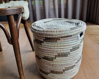May include: A round woven basket with a lid. The basket is made of natural fibers in shades of white, brown, and black. The basket is sitting on a wooden surface. The basket has a handle on each side.