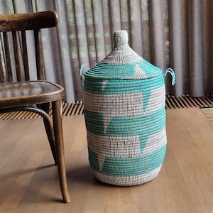 Op de afbeelding: Een cilindrische geweven mand met een deksel. De mand is turquoise en wit, met een herhalend geometrisch patroon. Hij heeft twee turquoise handvatten en een knop op het deksel. De mand staat op een houten vloer.