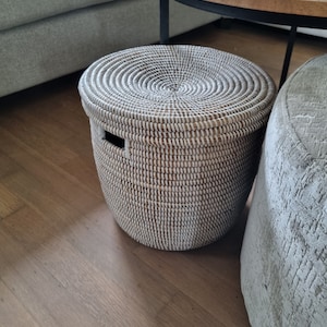 Op de afbeelding: Een ronde, geweven opbergmand met een deksel. De mand is wit en bruin, met een cirkelvormig patroon op het deksel. Hij heeft een handgreepuitsparing aan de zijkant. De mand staat op een houten vloer.
